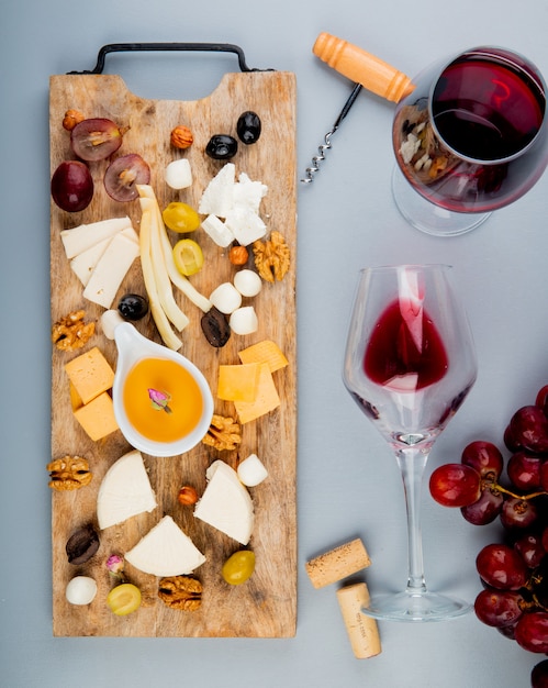 Vista dall'alto di burro con diversi tipi di formaggio uva olive noci sul tagliere e bicchieri di vino con tappi e cavatappi su bianco