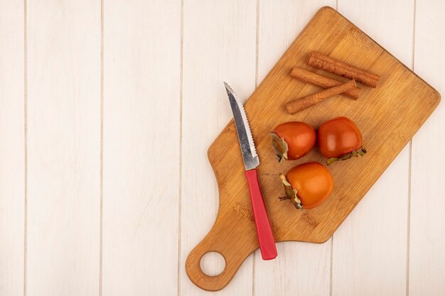 Vista dall'alto di cachi morbidi su una tavola da cucina in legno con bastoncini di cannella con coltello su una superficie di legno bianca con spazio di copia
