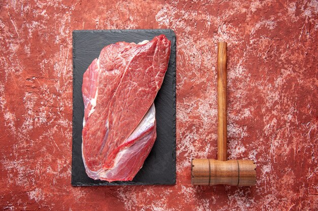 Vista dall'alto di carne fresca cruda rossa su tavola nera e martello di legno marrone su sfondo rosso pastello