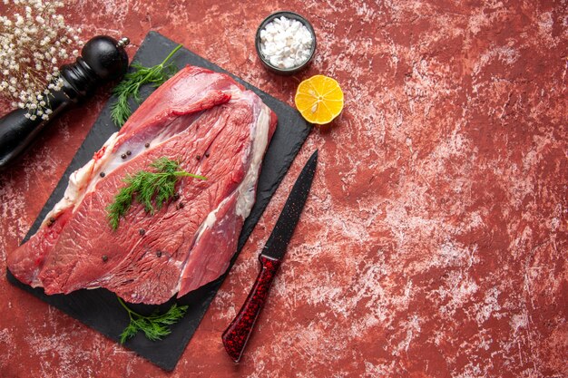 Vista dall'alto di carne rossa fresca cruda con verde e pepe su tavola nera coltello martello di legno sale limone su sfondo rosso pastello ad olio con spazio libero