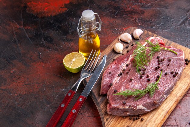 Vista dall'alto di carne rossa su tagliere di legno e forchetta e coltello di bottiglia di olio di pepe verde all'aglio su sfondo scuro