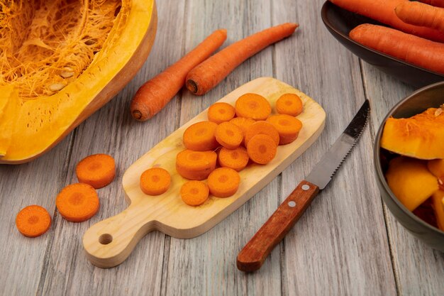 Vista dall'alto di carote tritate croccanti su una tavola da cucina in legno con coltello con carote su una ciotola con zucca isolata su un fondo di legno grigio