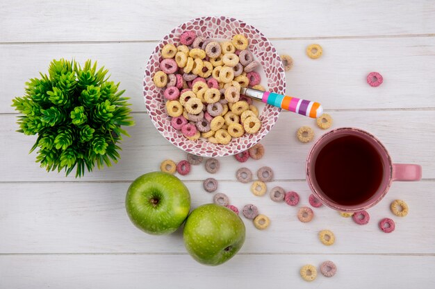 Vista dall'alto di cereali colorati sulla ciotola con cucchiaio colorato con una tazza di tè con mele verdi su bianco