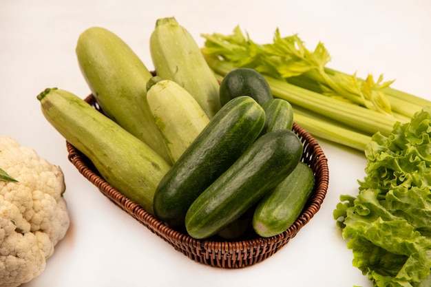 Vista dall'alto di cetrioli freschi e zucchine su un secchio con sedano cavolfiore e lattuga isolato su un muro bianco