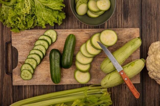 Vista dall'alto di cetrioli tritati e zucchine isolato su una tavola da cucina in legno con coltello con lattuga sedano e cavolfiore isolato su una parete in legno