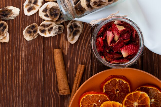 Vista dall'alto di chips di banane secche sparse da un barattolo di vetro e fette di fragola essiccate in un barattolo di vetro con fettine di arancia essiccate su un piatto su fondo in legno