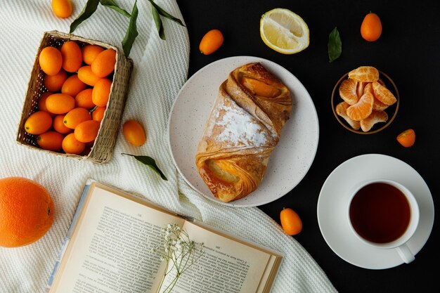 Vista dall'alto di croissant nel piatto e tazza di tè con fette di mandarino metà limone e libro aperto arancione con cesto di kumquat e fiorellino su libro su stoffa su sfondo nero