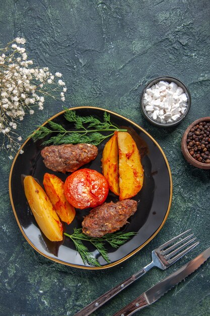 Vista dall'alto di deliziose cotolette di carne al forno con patate e pomodori su un piatto nero set di posate fiori bianchi spezie su sfondo verde nero colori misti