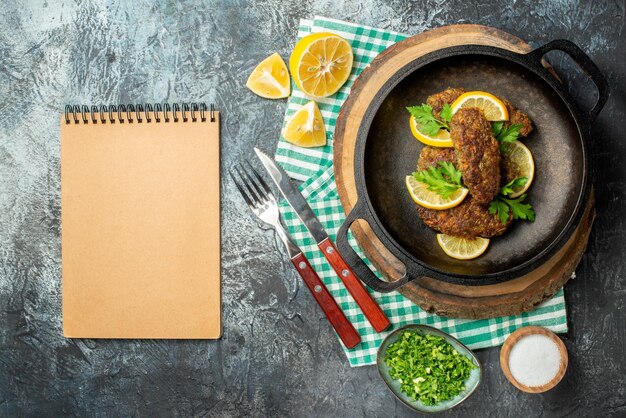 Vista dall'alto di deliziose cotolette servite con verdure e limone in padella nera su tavola di legno posate su asciugamano verde su sfondo scuro con spazio libero