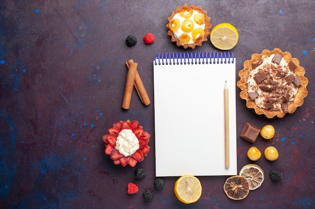 Vista dall'alto di deliziose torte con crema insieme a caramelle e blocco note del tè sulla superficie scura