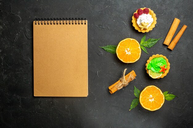 Vista dall'alto di deliziosi biscotti alla cannella lime e arance tagliate a metà con foglie e quaderno su sfondo scuro