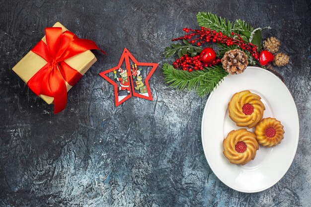 Vista dall'alto di deliziosi biscotti su un piatto bianco e decorazioni per il nuovo anno regalo con nastro rosso su superficie scura