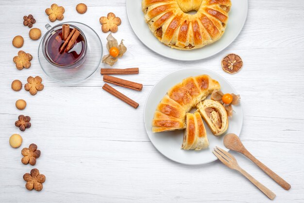 Vista dall'alto di deliziosi pasticcini a fette all'interno della piastra con ripieno insieme a tè alla cannella e biscotti su bianco, biscotto di pasticceria