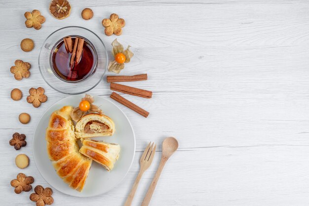 Vista dall'alto di deliziosi pasticcini affettati all'interno del piatto con ripieno insieme a tè e biscotti sulla scrivania bianca, biscotto di pasticceria dolce