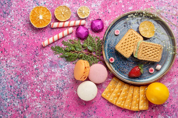 Vista dall'alto di deliziosi waffle con macarons francesi e cracker sulla scrivania rosa