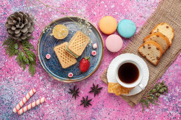 Vista dall'alto di deliziosi waffle con macarons francesi e tè sulla superficie rosa