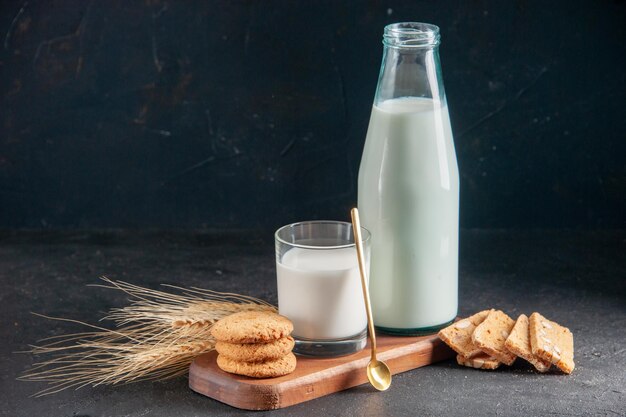 Vista dall'alto di delizioso latte in vetro e bottiglia di biscotti impilati cucchiaio d'oro su punte di vassoio di legno su superficie scura