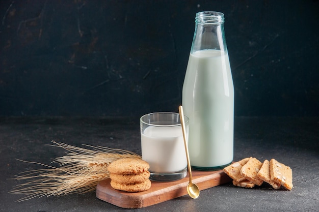 Vista dall'alto di delizioso latte in vetro e bottiglia di biscotti impilati cucchiaio d'oro su punte di vassoio di legno su superficie scura