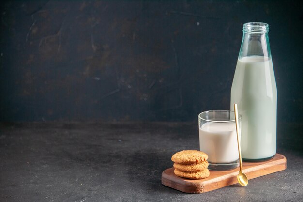 Vista dall'alto di delizioso latte in vetro e bottiglia di biscotti impilati cucchiaio d'oro sul vassoio di legno sul lato sinistro su sfondo scuro