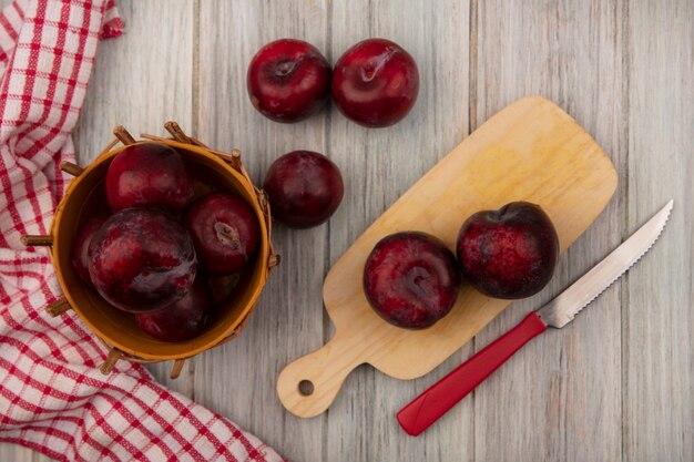 Vista dall'alto di dolci pluots isolato su una tavola da cucina in legno con coltello con pluots su un secchio su un panno controllato su una parete in legno grigio