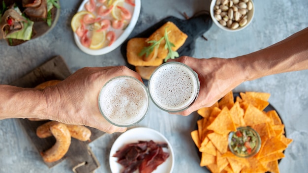 Vista dall'alto di due mani con bicchieri di birra e deliziosi snack.