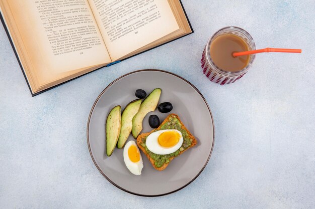 Vista dall'alto di fette di avocado fresco con avocado sulla fetta di pane con uovo imbustato e olive nere sulla piastra su bianco