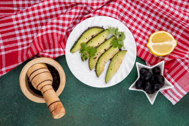 Vista dall'alto di fette di avocado fresco sulla piastra bianca con olive nere con mortaio di legno e pestello sulla tovaglia a quadretti rossa e verde