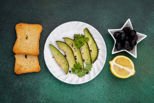 Vista dall'alto di fette di avocado sulla piastra bianca con olive nere fette di pane tostato e limone su gre