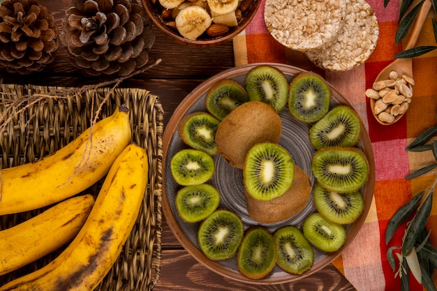 Vista dall'alto di fette di kiwi su un piatto e mazzo di banane in un cesto di vimini, cucchiaio di legno con arachidi e cracker di riso su legno