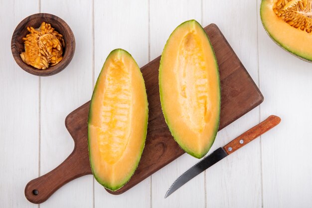 Vista dall'alto di fette di melone cantalupo fresco e delizioso sul bordo di cucina in legno con coltello con semi sulla ciotola di legno su legno bianco