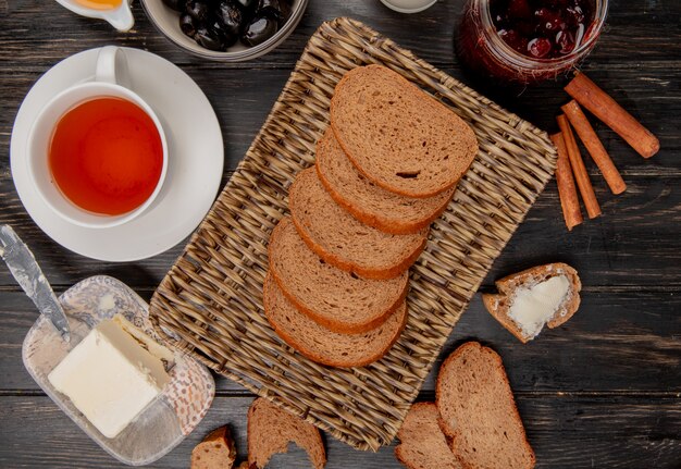 vista dall'alto di fette di pane di segale nel cestino piatto con una tazza di tè coltello burro marmellata di olive cannella sul tavolo di legno