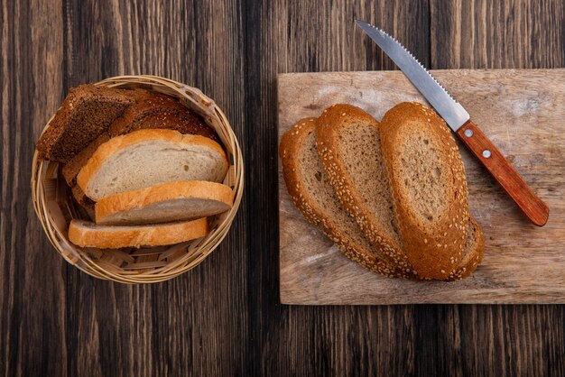 Vista dall'alto di fette di pane marrone pannocchia seminata con coltello sul tagliere e quelli bianchi di segale nel cestello su sfondo di legno