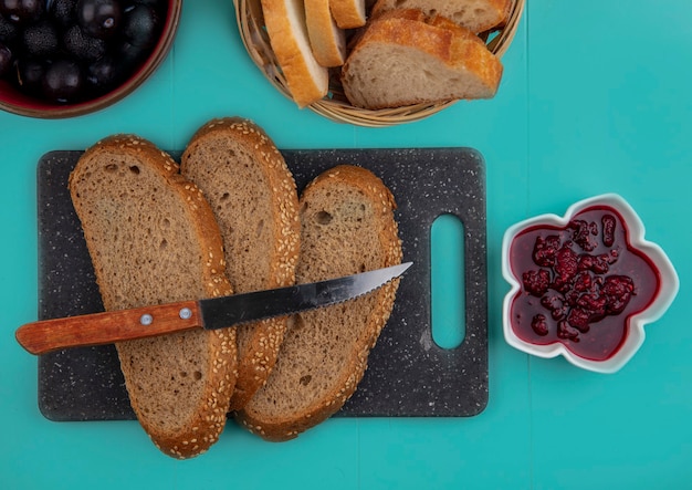 Vista dall'alto di fette di pannocchia marrone seminate con coltello sul tagliere e marmellata di lamponi con prugnole e fette di baguette su sfondo blu