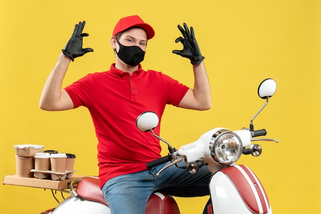 Vista dall'alto di fiducioso giovane adulto che indossa camicetta rossa e guanti cappello in maschera medica offrendo ordine seduto su uno scooter che spiega qualcosa su sfondo giallo