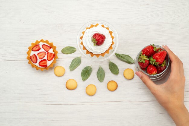 Vista dall'alto di fragole rosse fresche bacche dolci e deliziose con torte e biscotti sulla scrivania grigio chiaro, bacche di frutta rossa fresca