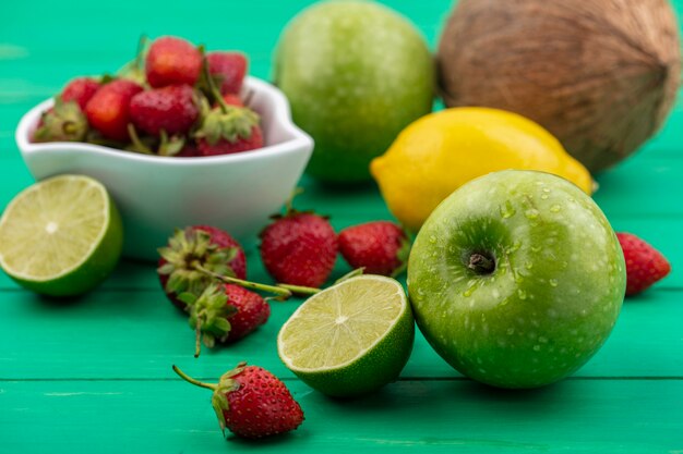 Vista dall'alto di fragole su una ciotola con frutta fresca come mele, limone e cocco isolato su uno sfondo verde