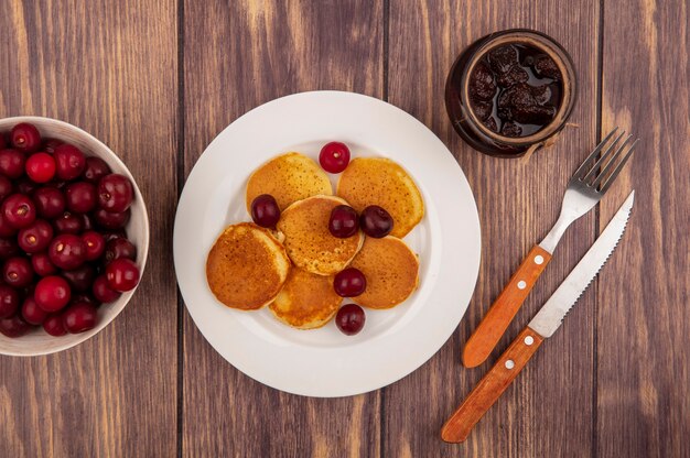 Vista dall'alto di frittelle con ciliegie nel piatto e ciotola di ciliegia con marmellata di fragole e coltello forchetta su fondo di legno