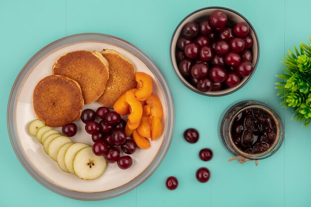 Vista dall'alto di frittelle con fette di albicocca e pera e ciliegie nel piatto con marmellata di fragole e ciliegie su sfondo blu