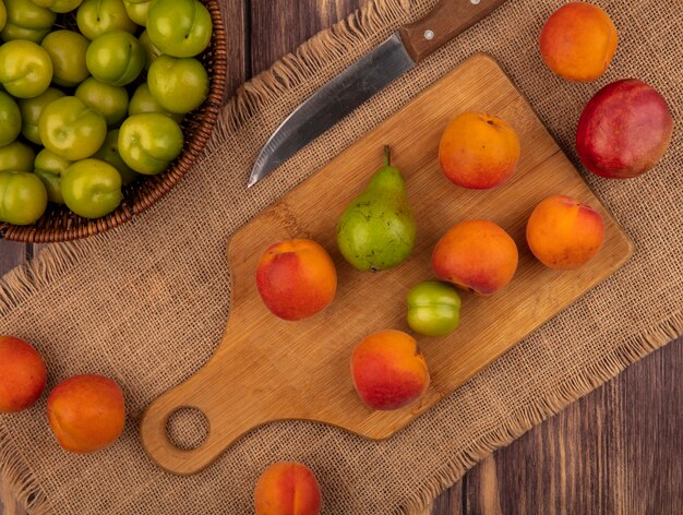 Vista dall'alto di frutta come albicocca pera prugna sul tagliere con pesca e coltello su tela di sacco e cesto di prugne su sfondo di legno