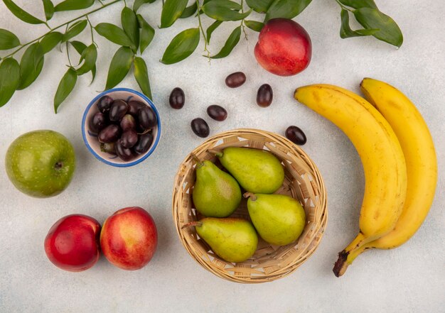 Vista dall'alto di frutta come cesto e ciotola di pera e uva con banana mela pesca con foglie su sfondo bianco