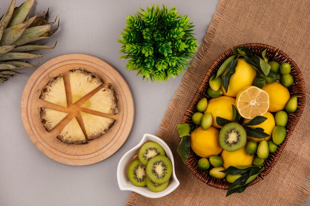 Vista dall'alto di frutta come kiwi kinkan e limoni su un secchio su un sacco di stoffa con fette di kiwi su una ciotola con fette di ananas su una tavola da cucina in legno su uno sfondo grigio