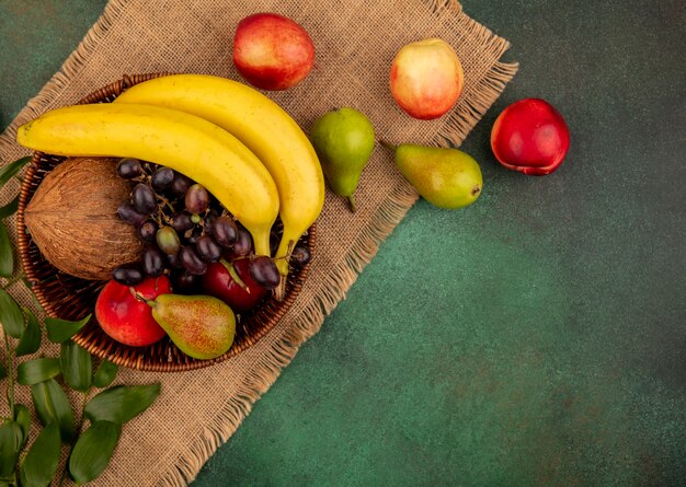 Vista dall'alto di frutta come noce di cocco banana uva pera pesca nel cestello e su tela di sacco su sfondo verde