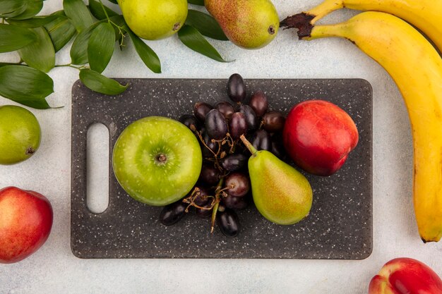 Vista dall'alto di frutta come pera mela uva pesca sul tagliere con banana e foglie su sfondo bianco