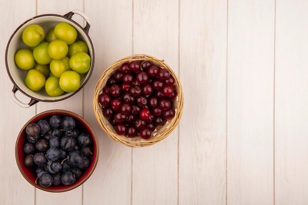 Vista dall'alto di frutta fresca come prugne ciliegia verdi su una ciotola con ciliegie rosse su un cesto con prugnole viola scuro su una ciotola rossa su uno sfondo di legno bianco con spazio di copia