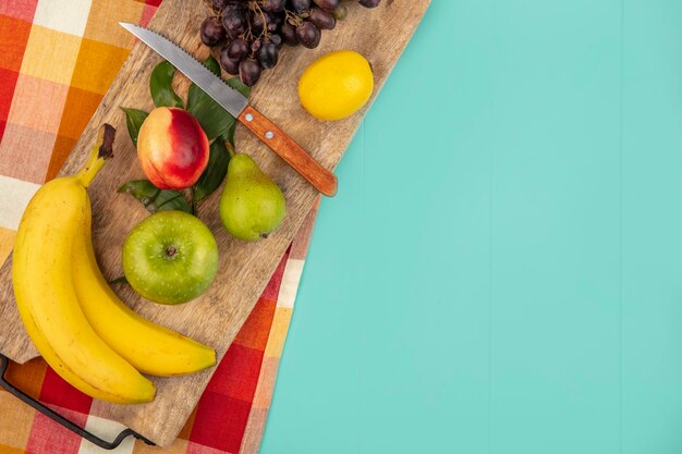 Vista dall'alto di frutti come banana mela pesca pera uva limone con coltello e foglie sul tagliere su panno plaid e sfondo blu con spazio di copia