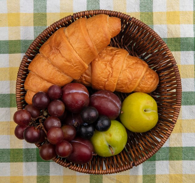 Vista dall'alto di frutti come uva pluots prugnole con croissant in cestino su sfondo di panno plaid