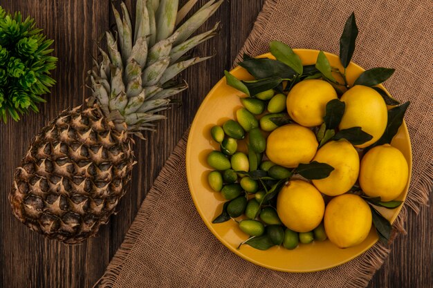 Vista dall'alto di frutti sani come limoni e kinkan su un piatto giallo su un panno di sacco con ananas isolato su una superficie in legno