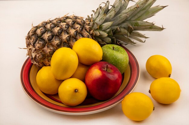 Vista dall'alto di frutti succosi come mele colorate di ananas e limoni su una ciotola con limoni isolato su un muro bianco