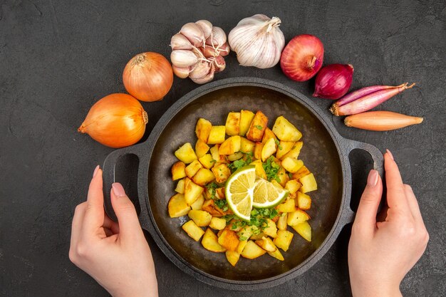 Vista dall'alto di gustose patate fritte all'interno della padella con limone e garlics intorno sulla superficie scura