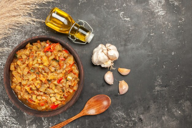 Vista dall'alto di insalata di melanzane arrosto in ciotola di olio bottiglia aglio cucchiaio di legno sulla superficie scura